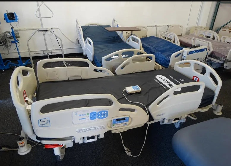 HillRomCareAssistEShospitalbedmodel Multiple hospital beds in a room with medical equipment.