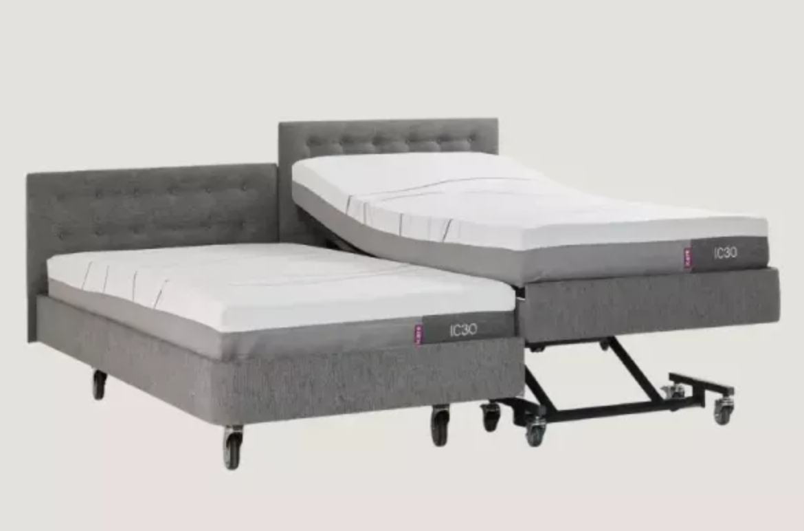 A dual adjustable bed frame system with two gray upholstered beds, one fully extended and the other partially raised to show the lift mechanism, on a light gray background.